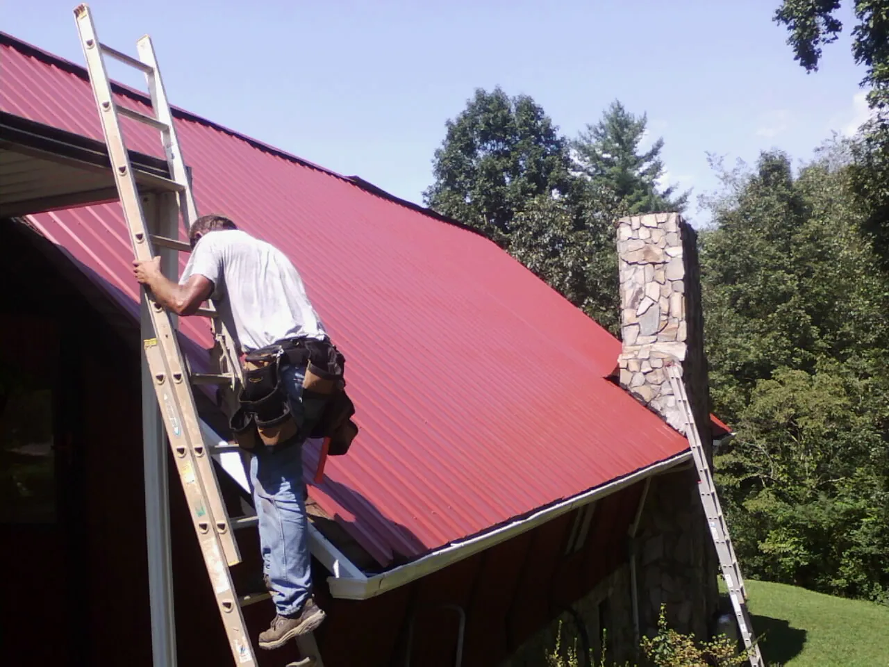 Roofer on a ladder beside a red metal roof and stone chimney — Soffit & Fascia Repair by Roofing Brothers in Tiptonville